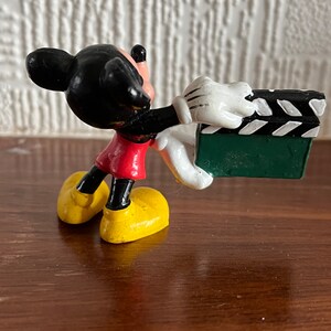 May include: A black and red Mickey Mouse figurine holding a green and white clapperboard. The figurine is made of plastic and has a cartoonish style.
