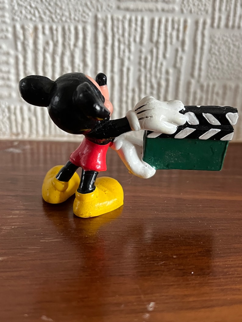 May include: A black and red Mickey Mouse figurine holding a green and white clapperboard. The figurine is made of plastic and has a cartoonish style.