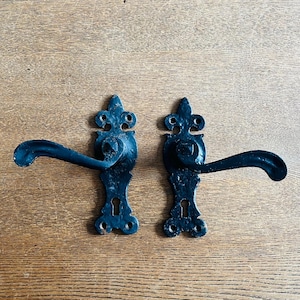 Peut inclure: Deux poignées de porte noires antiques avec des plaques arrière ornées de style fleur de lys. Les poignées ont un design incurvé et sont montées sur une surface en bois. La quincaillerie est probablement en fonte ou en métal similaire.
