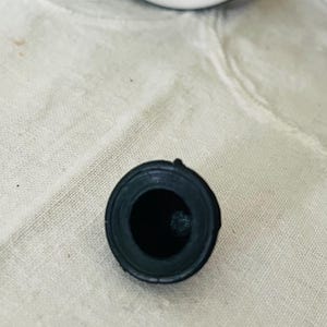 Op de afbeelding: Een zwarte flesstopper met een cirkelvormig ontwerp. De stopper heeft een donker interieur en een afgeronde buitenkant. De bovenkant van een zwart-witte fles is zichtbaar op de achtergrond. De stopper is waarschijnlijk gemaakt van rubber of een vergelijkbaar materiaal.