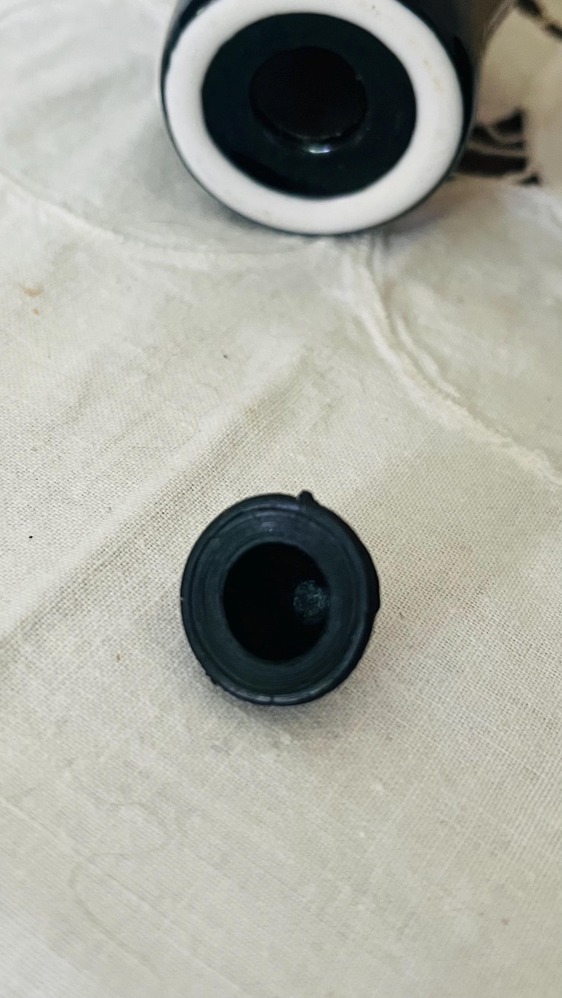 Op de afbeelding: Een zwarte flesstopper met een cirkelvormig ontwerp. De stopper heeft een donker interieur en een afgeronde buitenkant. De bovenkant van een zwart-witte fles is zichtbaar op de achtergrond. De stopper is waarschijnlijk gemaakt van rubber of een vergelijkbaar materiaal.