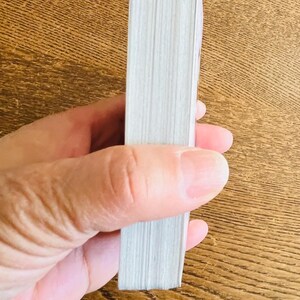 May include: A stack of white paper pages, likely from a book or notepad, held in a person's hand. The pages are tightly bound together, with a visible edge. The background is a wooden surface.