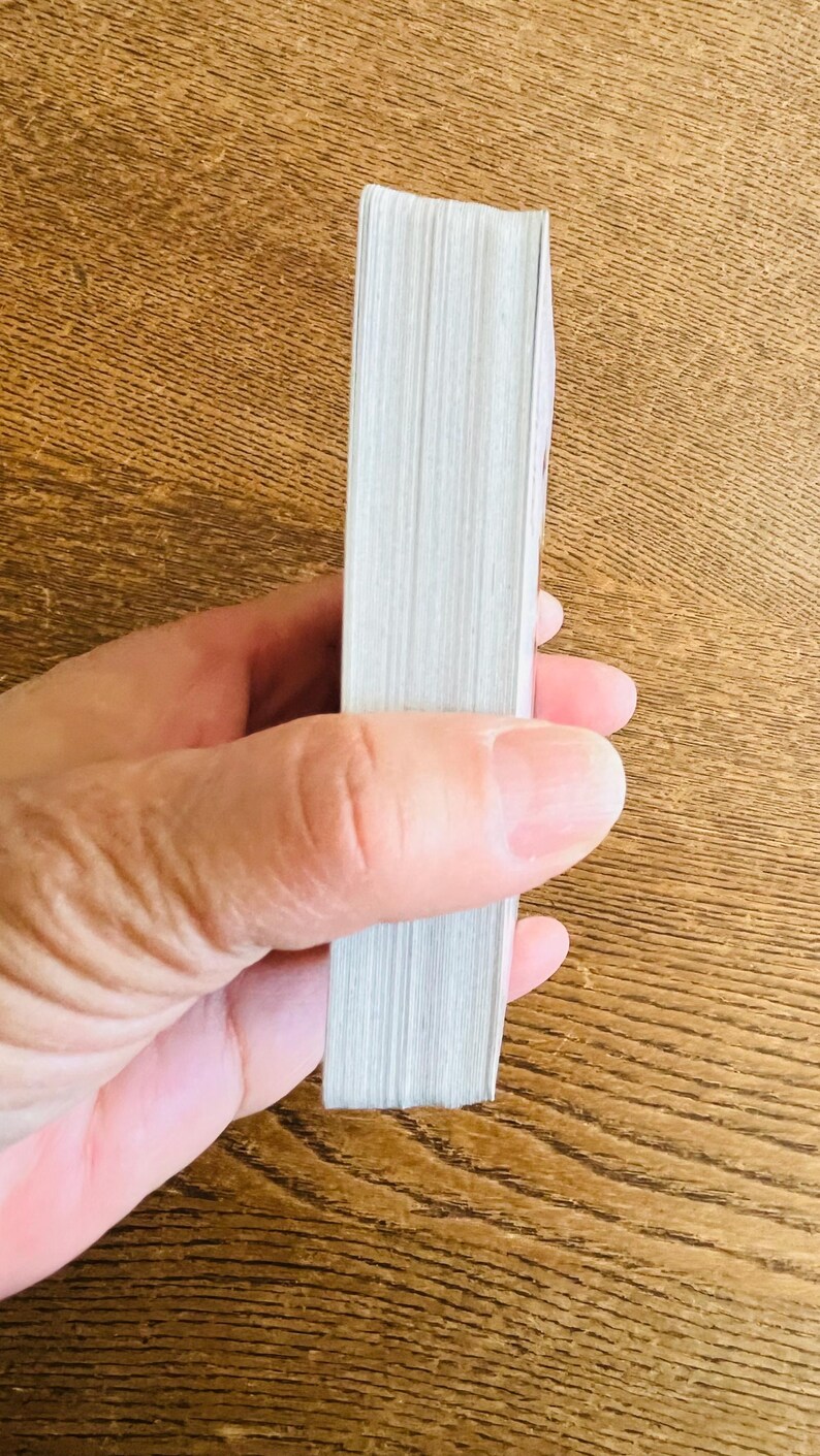 May include: A stack of white paper pages, likely from a book or notepad, held in a person's hand. The pages are tightly bound together, with a visible edge. The background is a wooden surface.