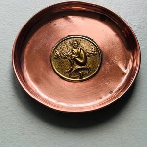 May include: A circular copper-coloured dish with a central, circular medallion. The medallion features a detailed, gold relief of a seated figure on a mushroom. The dish has a slightly raised edge and a polished surface.