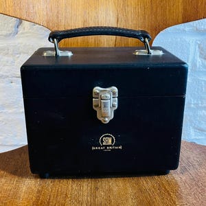 May include: A black vintage box with a handle and clasp, possibly for carrying items. The box has a small logo that reads "SFIM (GREAT BRITAIN)". The box is on a wooden chair.