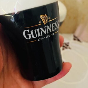 Op de afbeelding: Een zwart-wit Guinness pintglas met het Guinness logo in goud en wit. Het glas heeft een witte schuimkraag en het woord "Draught" staat onder het logo gedrukt. Het glas wordt in een hand gehouden.
