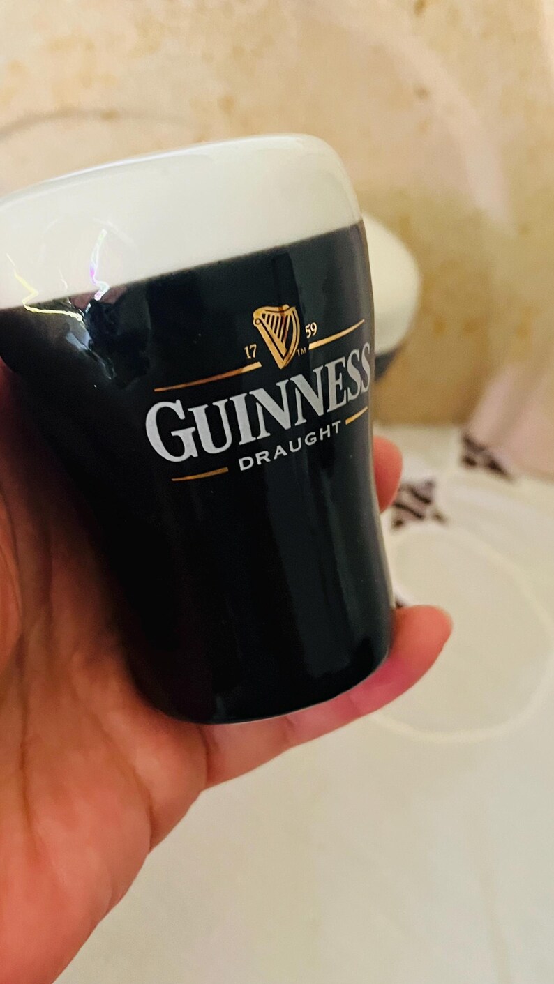 Op de afbeelding: Een zwart-wit Guinness pintglas met het Guinness logo in goud en wit. Het glas heeft een witte schuimkraag en het woord "Draught" staat onder het logo gedrukt. Het glas wordt in een hand gehouden.