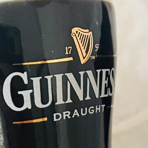 Op de afbeelding: Een close-up van een glas Guinness Draught. Het zwarte glas toont het gouden Guinness-logo, een harp, en de witte tekst 'Guinness Draught'. De drank is donker met een romige schuimkraag.