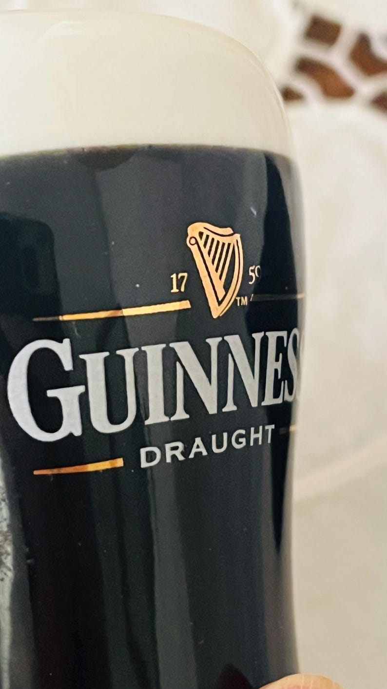 Op de afbeelding: Een close-up van een glas Guinness Draught. Het zwarte glas toont het gouden Guinness-logo, een harp, en de witte tekst 'Guinness Draught'. De drank is donker met een romige schuimkraag.