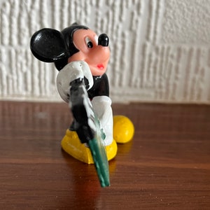 May include: A black and white plastic figurine of Mickey Mouse holding a green and white sign. Mickey is wearing yellow shoes and has a red mouth.