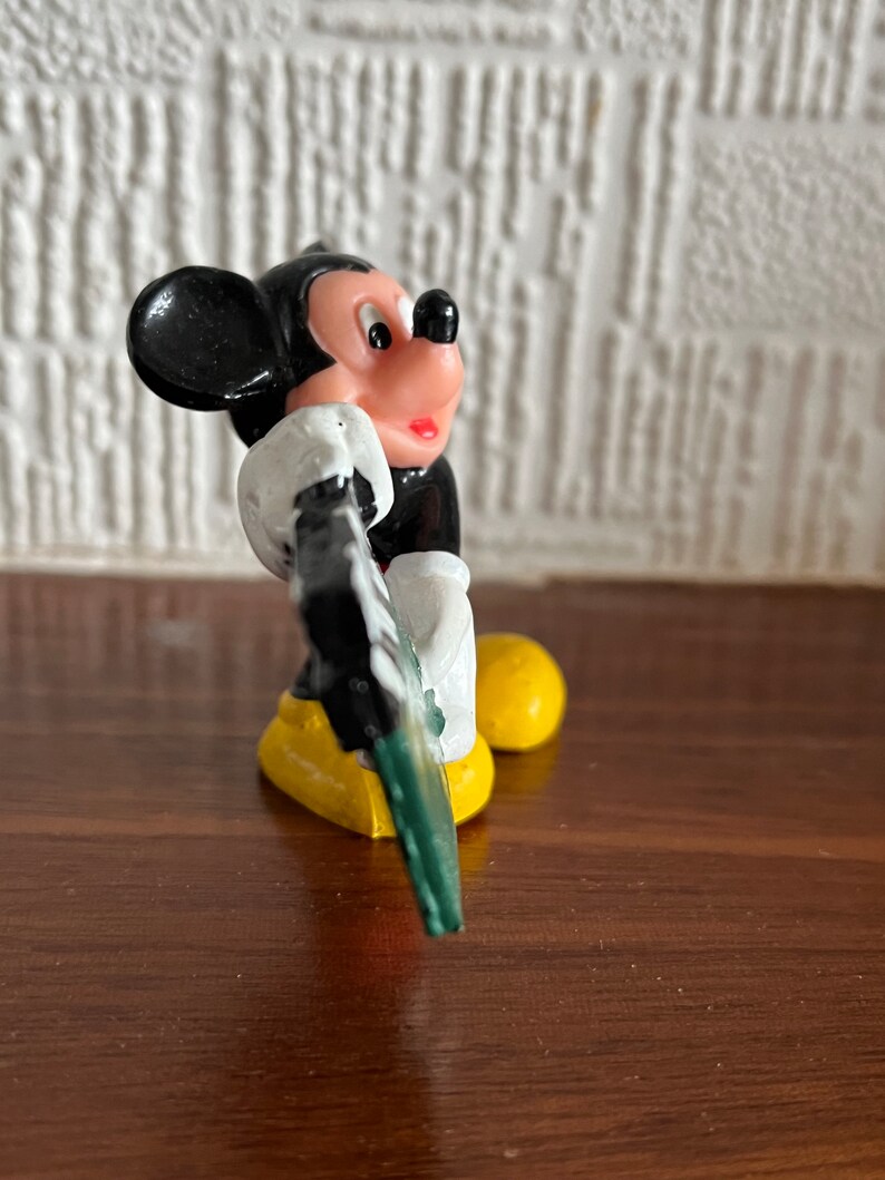 May include: A black and white plastic figurine of Mickey Mouse holding a green and white sign. Mickey is wearing yellow shoes and has a red mouth.