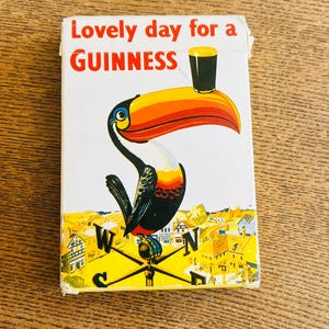 May include: Vintage Guinness advertisement featuring a toucan with a large orange and yellow beak, holding a pint of Guinness. The text "Lovely day for a Guinness" is displayed in red and white. The background shows a town.