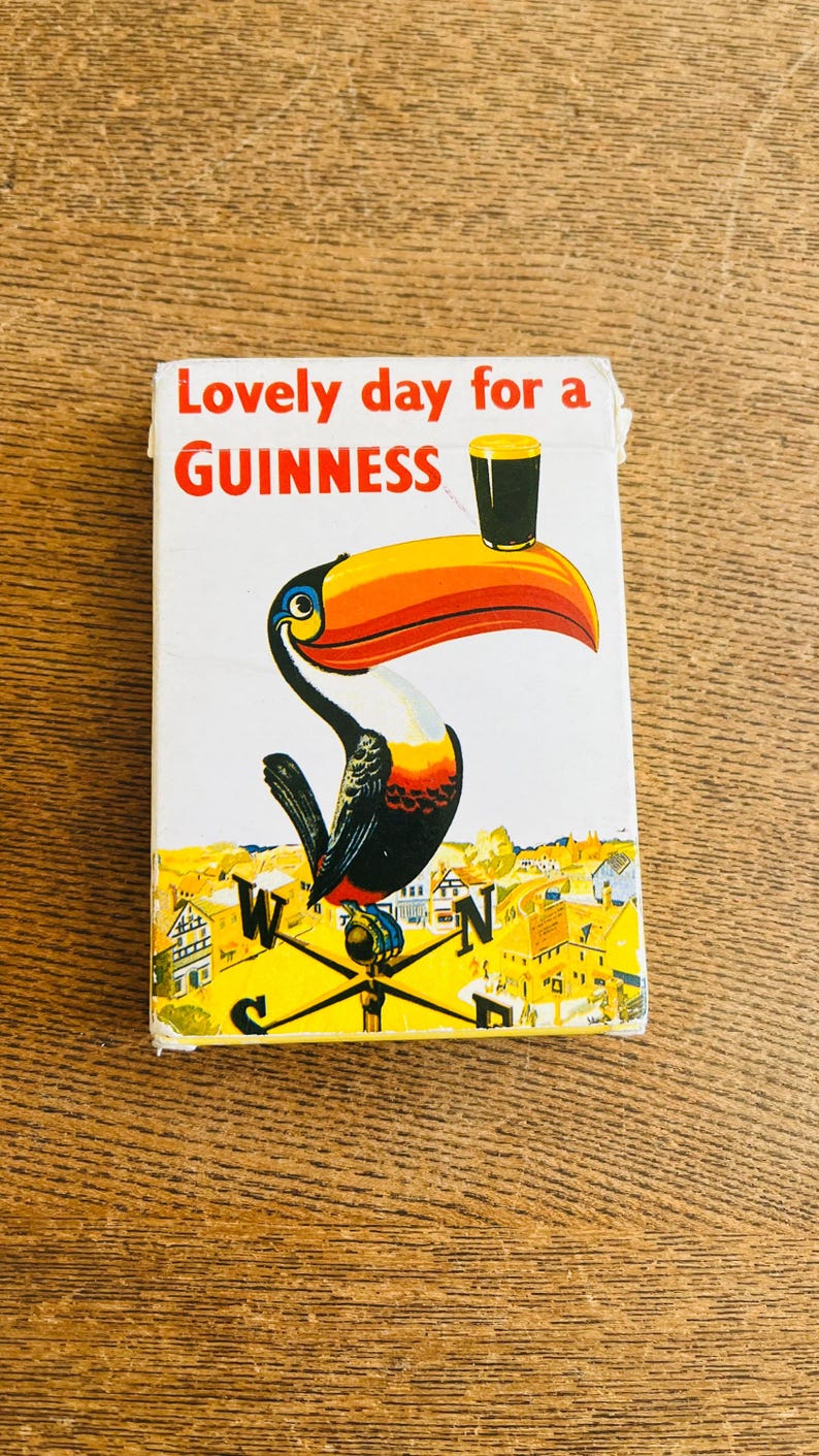 May include: Vintage Guinness advertisement featuring a toucan with a large orange and yellow beak, holding a pint of Guinness. The text "Lovely day for a Guinness" is displayed in red and white. The background shows a town.