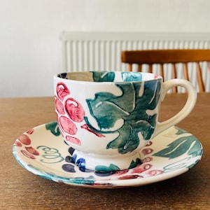May include: A white ceramic teacup and saucer set with a hand-painted grape design in red, blue, and green. The cup has a handle and sits on a matching saucer. The set is on a wooden table.