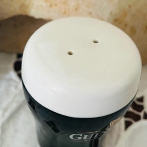 Op de afbeelding: Een donkergroene en witte keramische zoutstrooier. De zoutstrooier heeft een witte bovenkant met twee gaatjes en een donkergroene basis. Het woord "GU" is zichtbaar op de zijkant van de strooier.