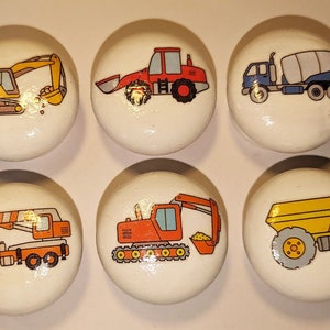 May include: Six white round wooden knobs with different construction vehicles painted on them. The vehicles include a yellow excavator, a red bulldozer, a blue cement mixer, an orange crane, an orange excavator, and a yellow dumper truck.