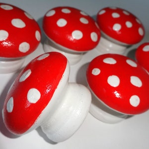 May include: Six mushroom-shaped decorative items. Each has a bright red cap with white spots and a white stem. The objects are arranged on a white surface, creating a whimsical display.
