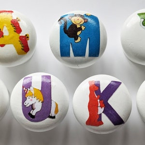 May include: Seven white wooden cabinet knobs featuring letters and animal illustrations. The letters are A, M, Y, L, U, K, and E. Animals include a lion, monkey, elephant, and unicorn. Ideal for children's furniture.