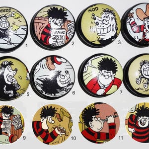 May include: A collection of twelve round cabinet knobs featuring colourful comic strip illustrations of characters from The Beano. Each knob displays a unique scene with black, red, and yellow ink details on a circular surface with a black base.