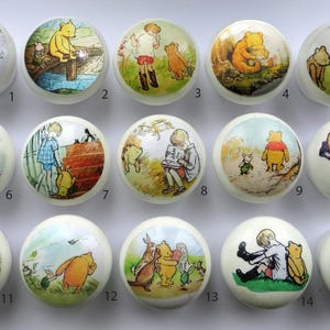 May include: A collection of fifteen round, white ceramic cabinet knobs, each featuring a unique, colourful illustration of Winnie the Pooh and friends. The knobs are arranged in three rows of five, with each knob numbered from one to fifteen.