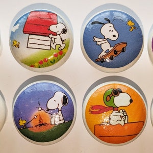May include: Set of eight round wooden knobs featuring different illustrations of Snoopy, the beloved beagle from Peanuts. Each knob has a different background colour and features Snoopy in a different pose or activity, including holding an umbrella, skateboarding, and flying a plane.