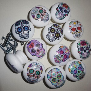 May include: Twelve white ceramic cabinet knobs with colourful sugar skull designs. Each knob features a different skull design with flowers and other decorative elements.