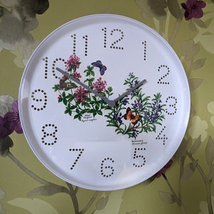 May include: White round clock with black numbers and hands. The clock face features a floral design with the words "Wild Thyme" and "Rosemary" printed on it.