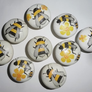 May include: Ten round, white painted stones with hand-painted bumblebees and yellow flowers. The bees have black and yellow stripes and grey wings.