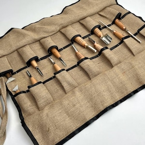 May include: A tan hessian tool roll featuring black trim and multiple pockets containing a set of wood carving tools. Each tool has a light-coloured wooden handle and a metallic blade of varying shapes, including gouges, chisels, and v-tools.