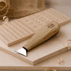 Op de afbeelding: Een houtsnijgereedschap met een lichtbruin houten handvat en een klein, driehoekig metalen mesje, rustend op een rechthoekige houten plank met ingewikkelde geometrische patronen, naast gladde houten planken.
