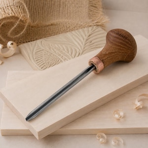 May include: A wood carving gouge with a dark metal blade and a rounded wooden handle featuring the engraved text STRYT. The tool is positioned on a light-colored wooden board, accompanied by small wood shavings and a carved wooden block.