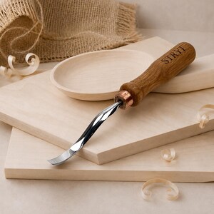 May include: A wood carving tool with a curved metal blade and a wooden handle featuring the brand name STRYI engraved on the side. The tool is positioned on a light-colored wooden board next to a small wooden bowl and scattered wood shavings.