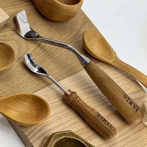 Puede incluir: Dos cuchillos de gancho de metal con hojas curvas y mangos de madera, cada uno con el nombre de la marca STRYI grabado en el mango. Las herramientas están dispuestas entre varios espacios en blanco de cucharas y cuencos de madera.