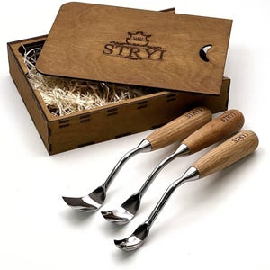 May include: Three metal wood carving tools with light brown wooden handles, each engraved with the word STRYI, arranged in front of an open wooden box containing wood shavings and a lid featuring the STRYI logo and a crown graphic.