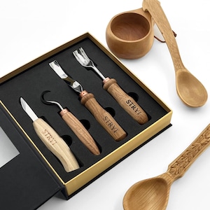 May include: A set of four wood carving tools with light wood handles branded with the text STRYI, housed in a black foam-lined box. Included are a straight knife, a hook knife, and two gouges. Also shown are a wooden cup and a carved wooden spoon.