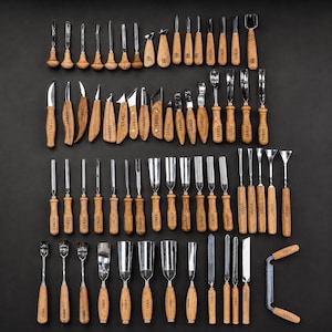 May include: A large collection of wood carving tools with light brown wooden handles and silver metal blades. Each handle is branded with the text STRYI. The set includes various gouges, chisels, knives, and a drawknife, all arranged in rows.
