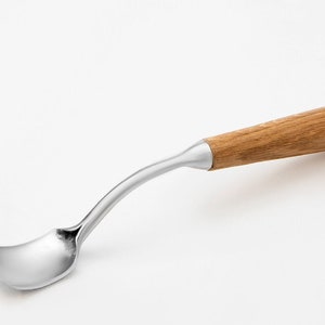 以下が含まれることがあります： 曲がった柄と木製のグリップが付いたステンレス製のスプーン。柄は明るい茶色の木材でできており、「STRYI」の文字が刻まれています。スプーンは、すくうように曲がったボウル型をしています。