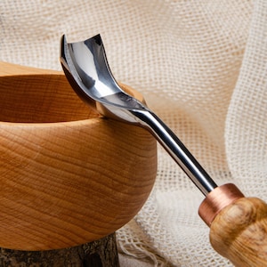 May include: A close-up of a wood carving tool with a curved blade and a wooden handle. The tool is resting on a wooden bowl.
