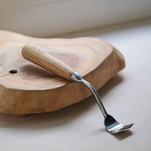 May include: A wood carving tool with a light brown wooden handle featuring the engraved text STRYT and a curved, polished metal blade. The tool has a cylindrical handle that tapers into a metallic neck, connecting to a scoop-shaped metal tip.
