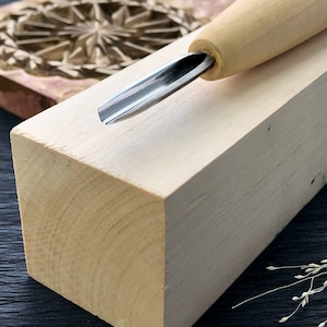 以下が含まれることがあります： 木製のハンドルと金属製の刃を持つ木彫りの道具のクローズアップ。道具は木製のブロックの上に置かれています。