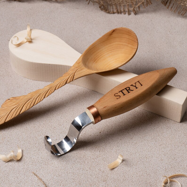 May include: A wooden spoon with a carved design and a wooden carving knife with a metal blade and a copper ferrule. The knife has the word "STRYI" engraved on the handle.