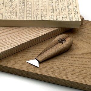 Op de afbeelding: Een houtsnijgereedschap met een driehoekig metalen blad en een gladde, lichtbruine houten handgreep met een klein, donker cirkelvormig logo. Het gereedschap ligt op een lichtgekleurde houten plank met ingegraveerde geometrische patronen.