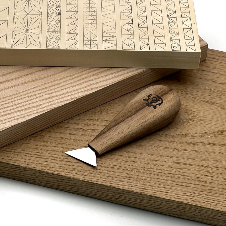 Op de afbeelding: Een houtsnijgereedschap met een driehoekig metalen blad en een gladde, lichtbruine houten handgreep met een klein, donker cirkelvormig logo. Het gereedschap ligt op een lichtgekleurde houten plank met ingegraveerde geometrische patronen.