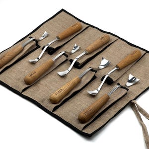 Peut inclure: Un ensemble de six outils de sculpture sur bois avec des manches en bois brun clair et des lames en m&eacute;tal argent&eacute;, chacun marqu&eacute; du texte STRYI. Les outils sont dispos&eacute;s dans un organisateur en tissu beige avec bordure noire.