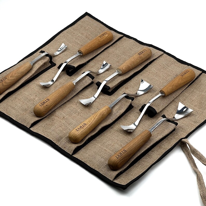Peut inclure: Un ensemble de six outils de sculpture sur bois avec des manches en bois brun clair et des lames en m&eacute;tal argent&eacute;, chacun marqu&eacute; du texte STRYI. Les outils sont dispos&eacute;s dans un organisateur en tissu beige avec bordure noire.