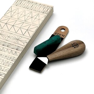 Op de afbeelding: Een lichtgekleurde houten plank met geometrische lijnpatronen, een snijgereedschap met houten handvat en metalen mes, een zwarte leren aanzetriem en een rechthoekig groen polijstblok. Het handvat heeft een kleine logo-gravure.