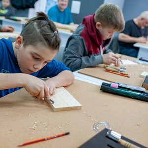 Op de afbeelding: Een lichtgekleurd rechthoekig houten blok dat wordt bewerkt met een metalen guts. De werkplek bevat een potlood, een set snijgereedschap in een zwarte schuimrubberen organizer en houtkrullen verspreid over het bruine oppervlak tijdens het werk.