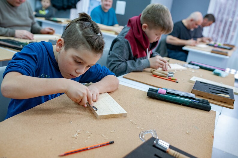 Op de afbeelding: Een lichtgekleurd rechthoekig houten blok dat wordt bewerkt met een metalen guts. De werkplek bevat een potlood, een set snijgereedschap in een zwarte schuimrubberen organizer en houtkrullen verspreid over het bruine oppervlak tijdens het werk.