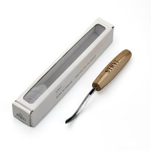 Op de afbeelding: Een haakmes voor houtsnijwerk met een gebogen metalen blad en een lichtbruin houten handvat met de merknaam STRYI. Het gereedschap ligt naast de witte rechthoekige kartonnen verpakking met de tekst STRYI BORN IN UKRAINE.