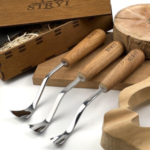 May include: Three metal wood carving tools with curved blades and light brown wooden handles, each branded with the text STRYI. The tools are arranged near a wooden box with the STRYI logo, a wooden block, and a carved wooden piece.